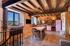 Una cocina rústica con vigas de madera a la vista, un comedor con mesa y sillas, y vistas a través de una ventana.