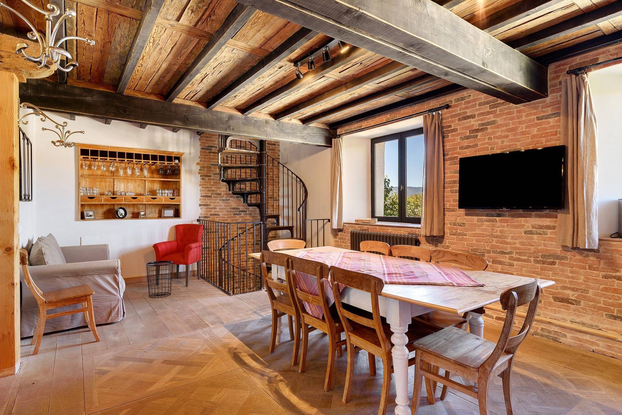 Un intérieur rustique avec des poutres apparentes, un mur de briques, une table à manger avec des chaises, une chaise rouge, un escalier et un téléviseur fixé au mur.