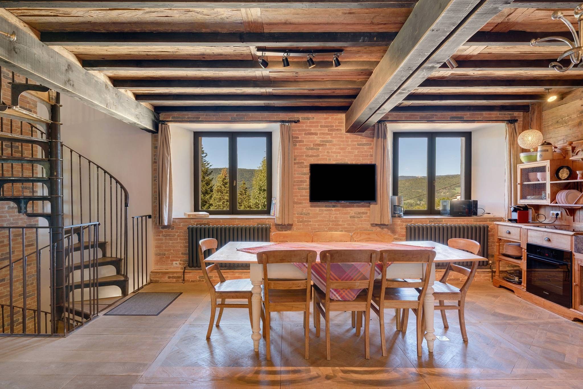 Un comedor con pared de ladrillo, vigas de madera, una mesa con sillas y un televisor montado en la pared.