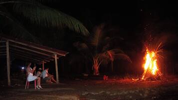 A group of people sitting under a shelter with a fire burning in the background.