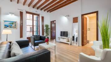 Amplia sala de estar con un sofá de cuero negro, una mesa de centro blanca y un televisor. Cuenta con techo de madera y una puerta que da a un baño.
