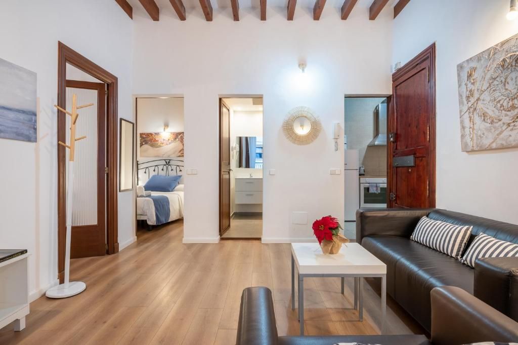 Una sala moderna con un sofá, una mesa de centro y un banco. Se ve una puerta que lleva a un dormitorio con una cama y un baño con un espejo y un lavabo.