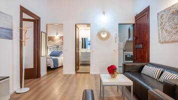 Una sala moderna con un sofá, una mesa de centro y un banco. Se ve una puerta que lleva a un dormitorio con una cama y un baño con un espejo y un lavabo.