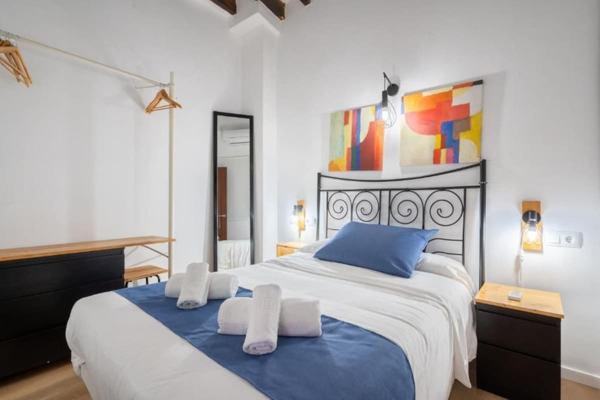 A hotel room with a bed, a bedside table, a mirror, and a wall with abstract art.
