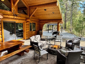 Una cabaña de troncos con un patio que incluye mobiliario exterior, una fogata y vistas del bosque circundante.