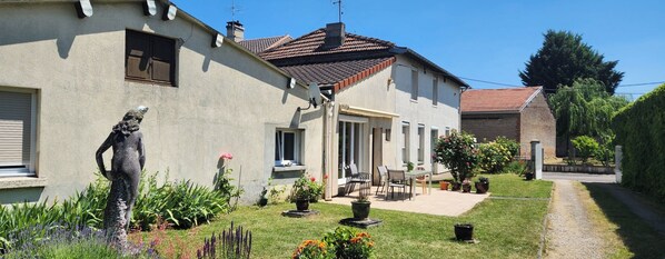 Een tweelaags huis met een terras, buitenmeubilair en een tuin met verschillende planten en bloemen.