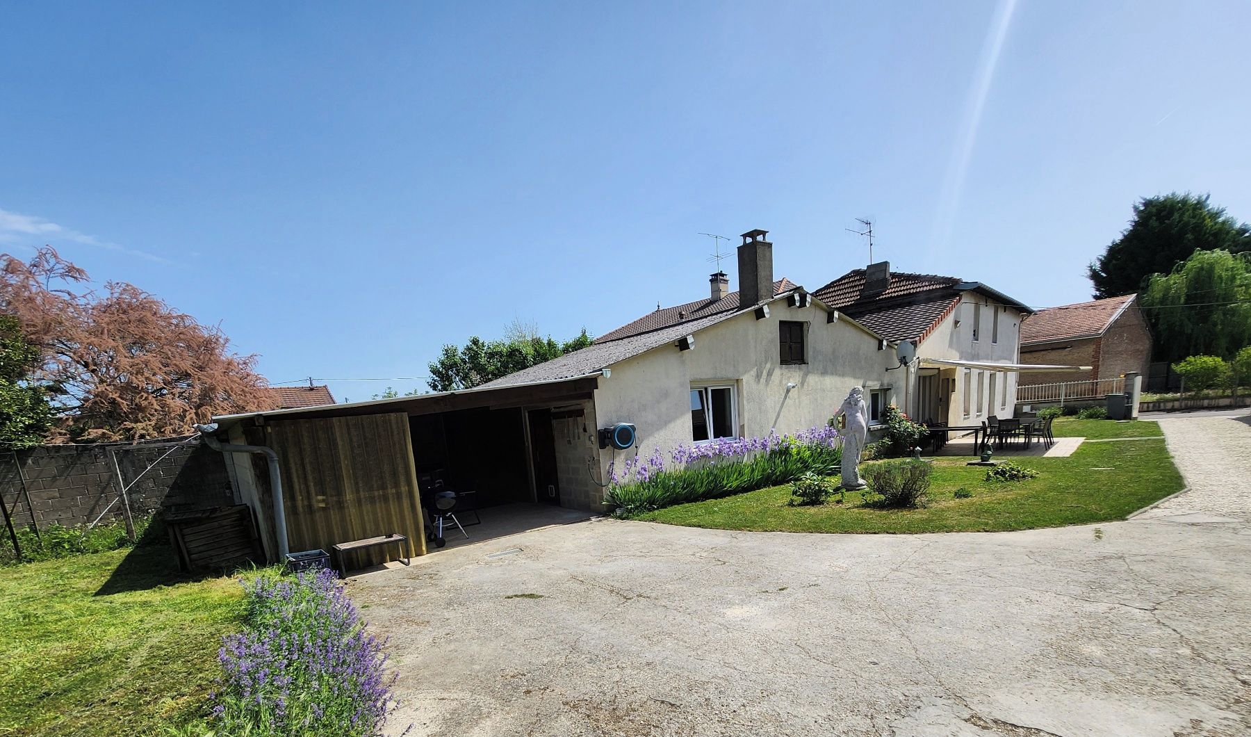 Una casa de dos pisos con garaje, un pequeño jardín con flores moradas y un cielo despejado.