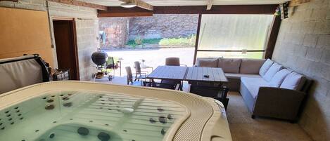 A hot tub in a covered outdoor area with a table and chairs, a sofa, and a view of the outdoors.