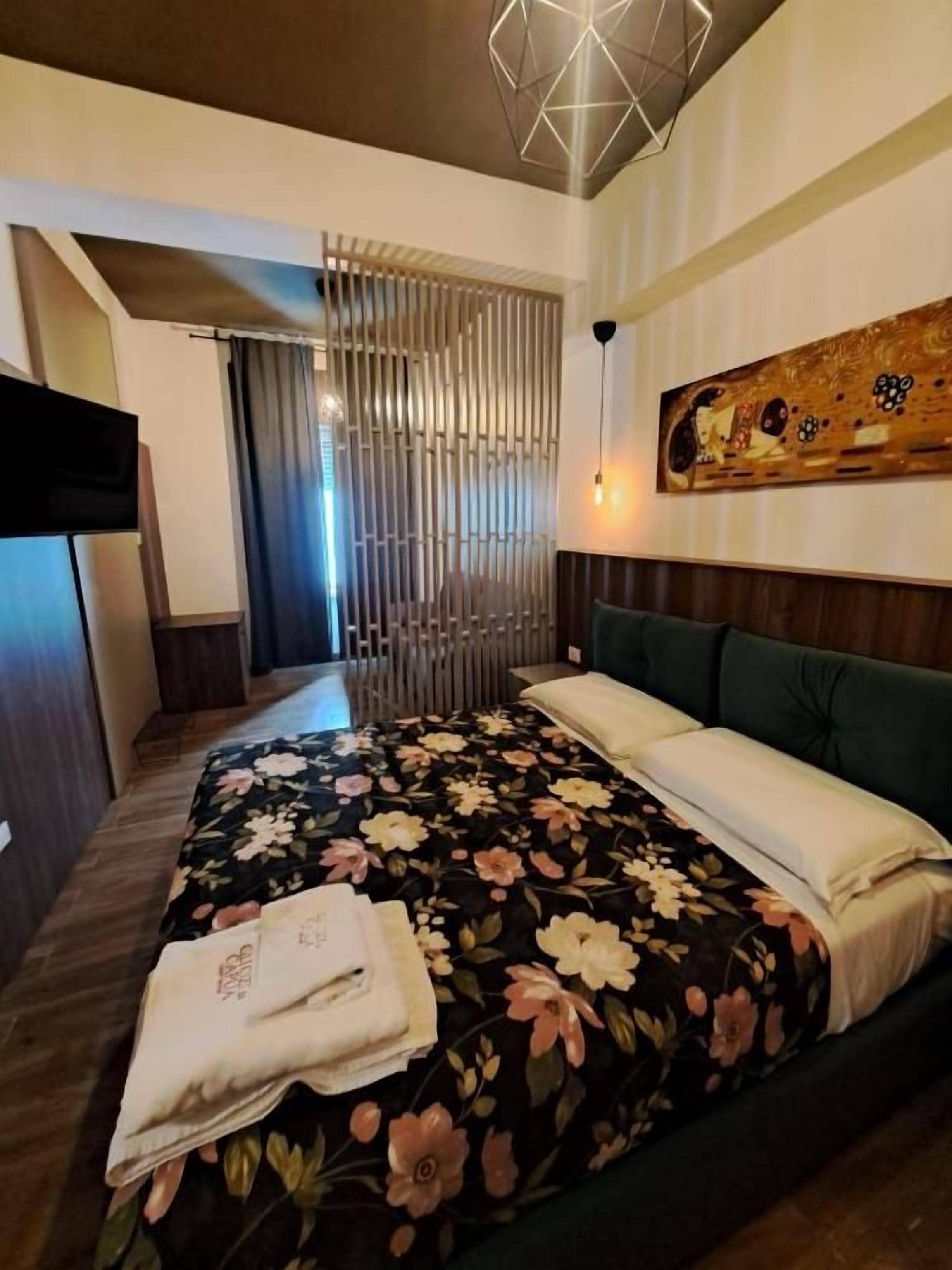 A hotel room with a bed, a television, a painting, and a modern light fixture.