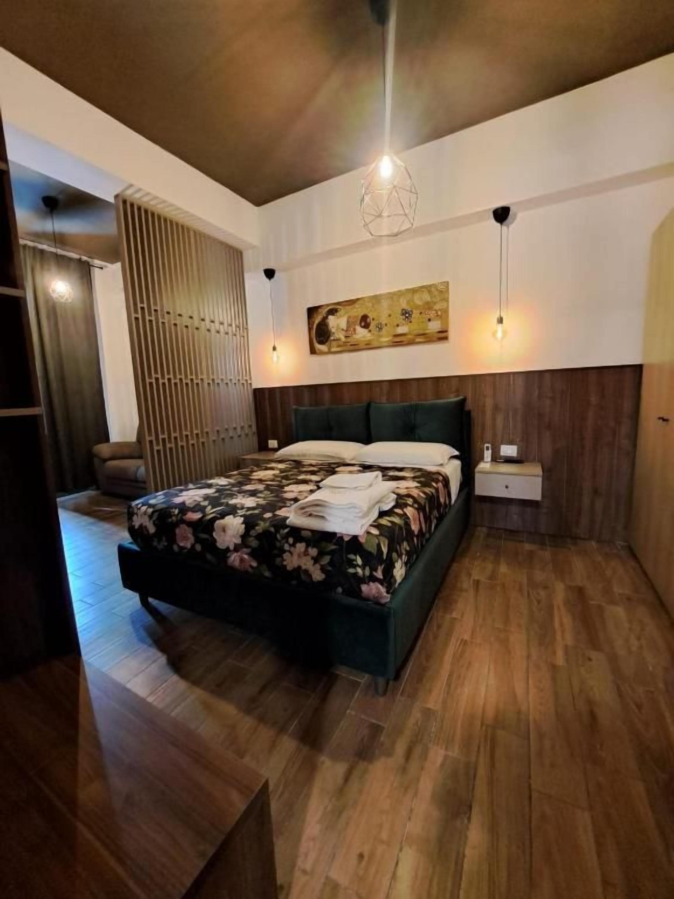 A modern bedroom with a large bed, wooden flooring, a wall-mounted artwork, and a hanging light fixture.