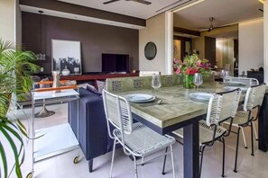 A dining area with a table set for four, surrounded by chairs, a television, and a decorative vase with flowers. - Pier 57 511 | Rooftop Pool Retreat (Puerto Vallarta)