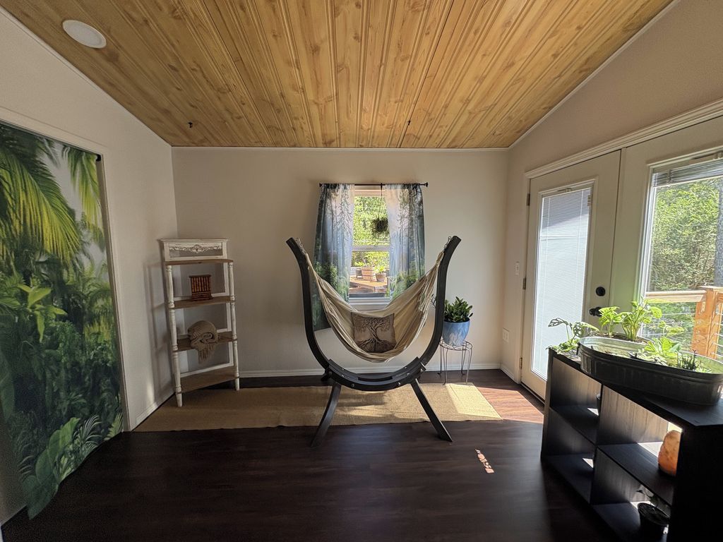 A room with a hammock chair, a wooden ceiling, a large window, and a plant shelf.