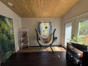 Una habitación con una silla hamaca, techo de madera, un ventanal grande y una estantería con plantas.