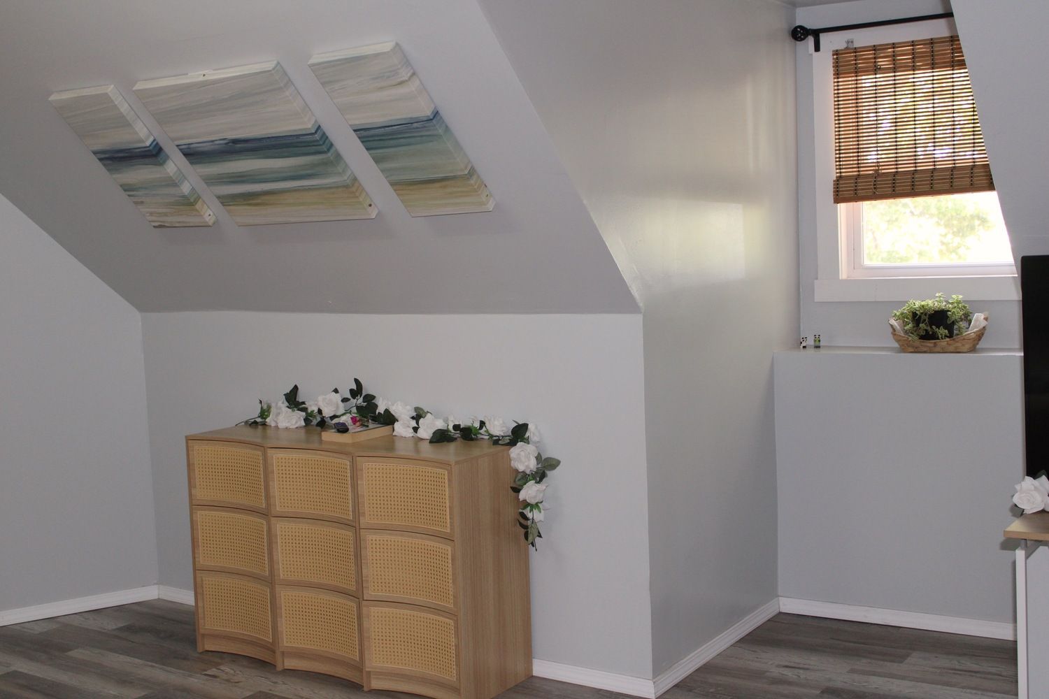 A room with a sloped ceiling, a rattan cabinet, a window with blinds, and a small plant on the windowsill.