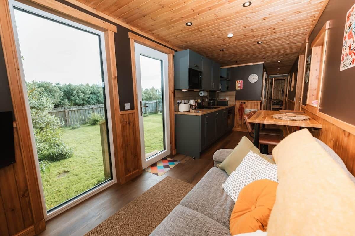 A cozy interior with a wooden ceiling, a sofa with cushions, a kitchen area, and a sliding glass door leading to a garden.
