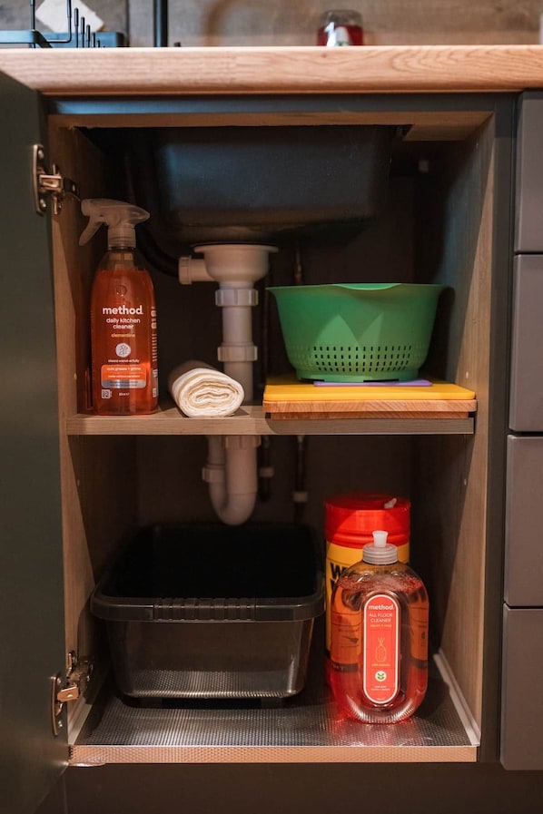 A kitchen cabinet with a built-in sink, a green colander, a bottle of Method all floor cleaner, and a black trash bin.