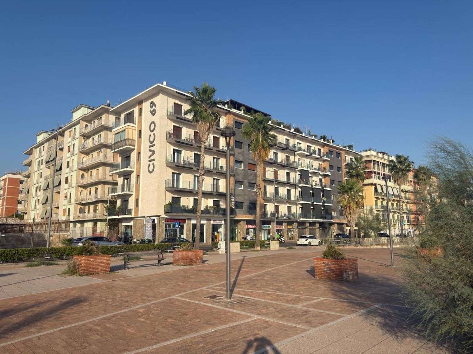 A multi-story residential building with balconies, a paved courtyard, and surrounding greenery.