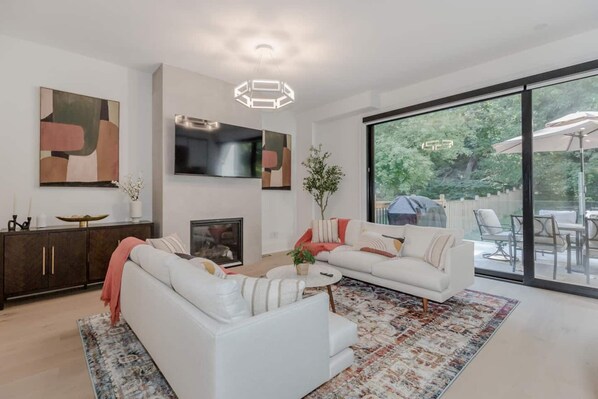 Un salon moderne avec un canapé blanc, une table basse et une cheminée. Une grande fenêtre offre une vue sur l’extérieur.