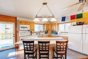 Una cocina con armarios de madera, una isla central con sillas, microondas, refrigerador y ventilador de techo.