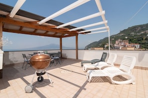 Eine überdachte Terrasse im Freien mit einem Grill, Liegestühlen und Blick auf eine gebirgige Landschaft.