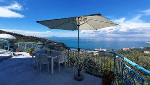 Eine Terrasse mit Tisch und Stühlen, einem großen Sonnenschirm und Blick auf das Meer und die Ferne.