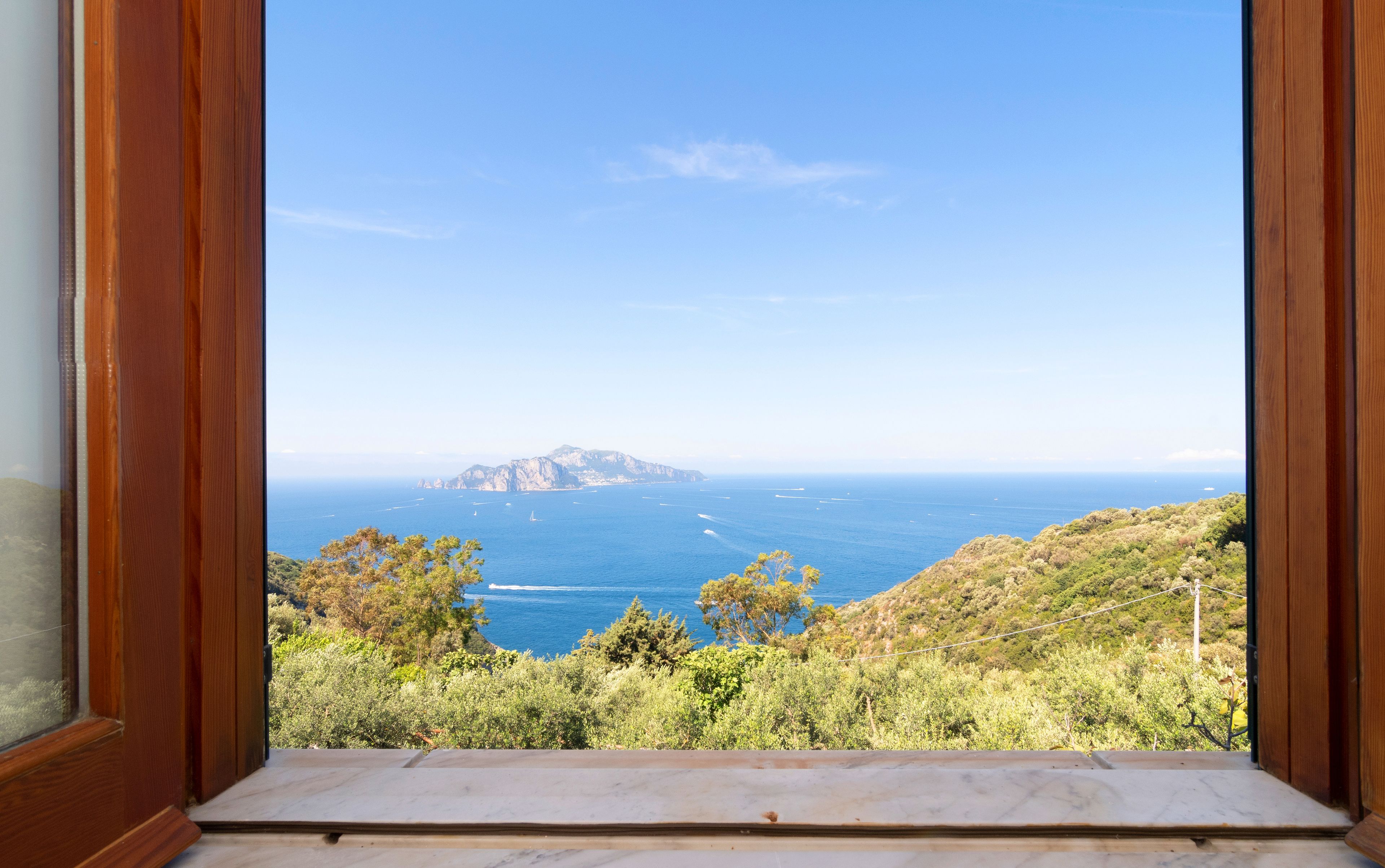 Une vue à travers une fenêtre à cadre en bois, donnant sur un paysage côtier avec un ciel dégagé, une terre lointaine et une végétation luxuriante.