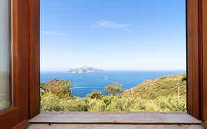 A view through a wooden-framed window overlooking a coastal landscape with a clear sky, distant landmass, and greenery. - Essences of the Mediterranean by Espresso Rentals (Termini)