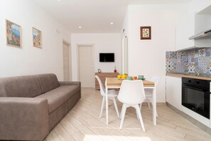 A modern living space with a sofa, a dining table with chairs, a kitchen area with a stove and oven, and a TV mounted on the wall. - Nonna Mary - Limone by Espresso Rentals (Sant'Agata Sui Due Golfi)