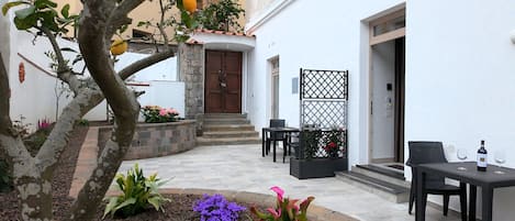 A courtyard with a stone bench, a table with a bottle and glasses, and a door leading to a building.