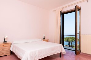 Ein Schlafzimmer mit einem Bett, zwei Nachttischen, einem Fenster mit Meerblick und einer Tür, die auf einen Außenbalkon führt.
