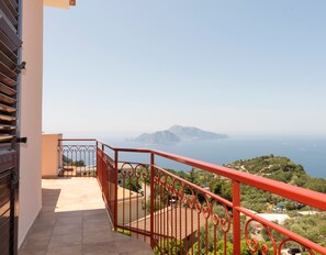 Varanda com guarda-corpo vermelho com vista para a paisagem costeira, montanhas e o mar.