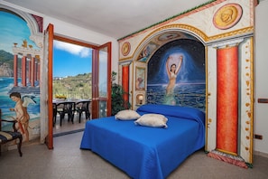Een slaapkamer met een bed, uitzicht op zee en een muurschildering van een vrouw in het water.