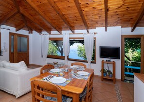 A cozy room with a wooden ceiling, a dining table set for four, a sofa, a TV, and a view of the sea. - Vitamin Sea by Espresso Rentals (Napoli)