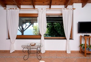 A room with a large window, white curtains, a small table with a basket, and a television on a wooden stand. - Vitamin Sea by Espresso Rentals (Napoli)