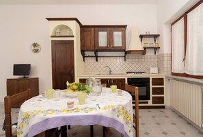 A compact kitchen with wooden cabinets, a dining table with a floral tablecloth, and a small refrigerator. - Limoneto - Combo by Espresso Rentals (Massa Lubrense)