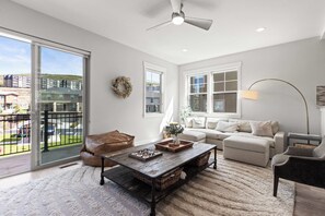 A spacious living room with a large sliding glass door to a balcony, a ceiling fan, a wooden coffee table, a beige sofa, and a floor lamp. - Gone Ski Inn! Cozy Townhome Perfect for Families (Kamas)