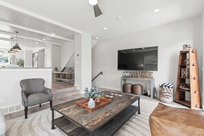 A modern living room with a large flat-screen TV, a fireplace, a staircase, and a cozy seating area. - Gone Ski Inn! Cozy Townhome Perfect for Families (Kamas)