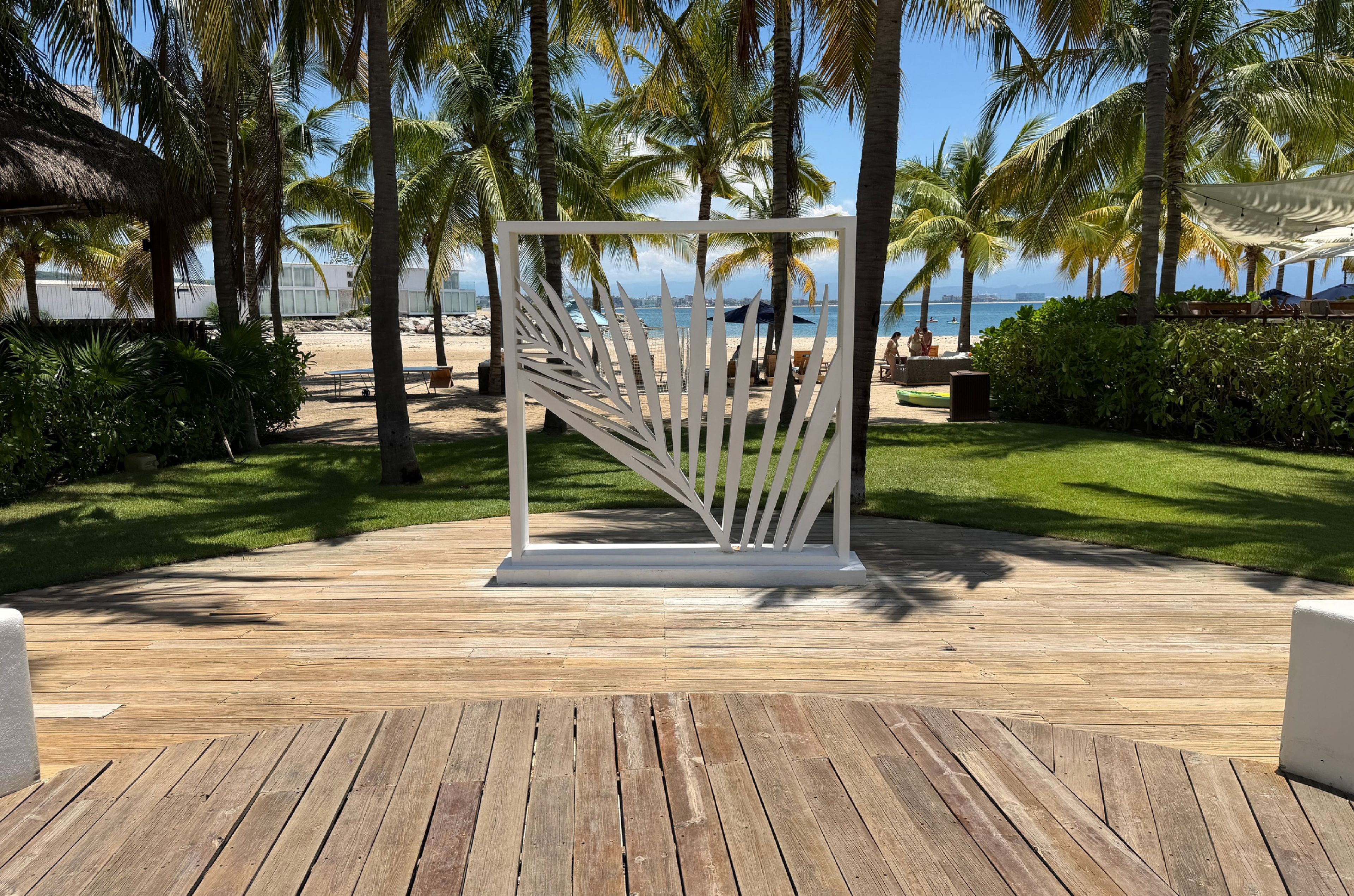Een houten terras met een witte decoratieve constructie, omgeven door palmbomen en uitzicht op het strand.