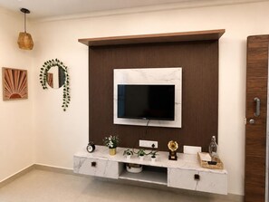 A modern living room with a flat-screen TV mounted on a wooden panel wall, a marble console table with decorative items, and a door on the right. - Leafy Boho 1BHK in HSR Layout, near Koramangala,AC,WiFi (Bengaluru)