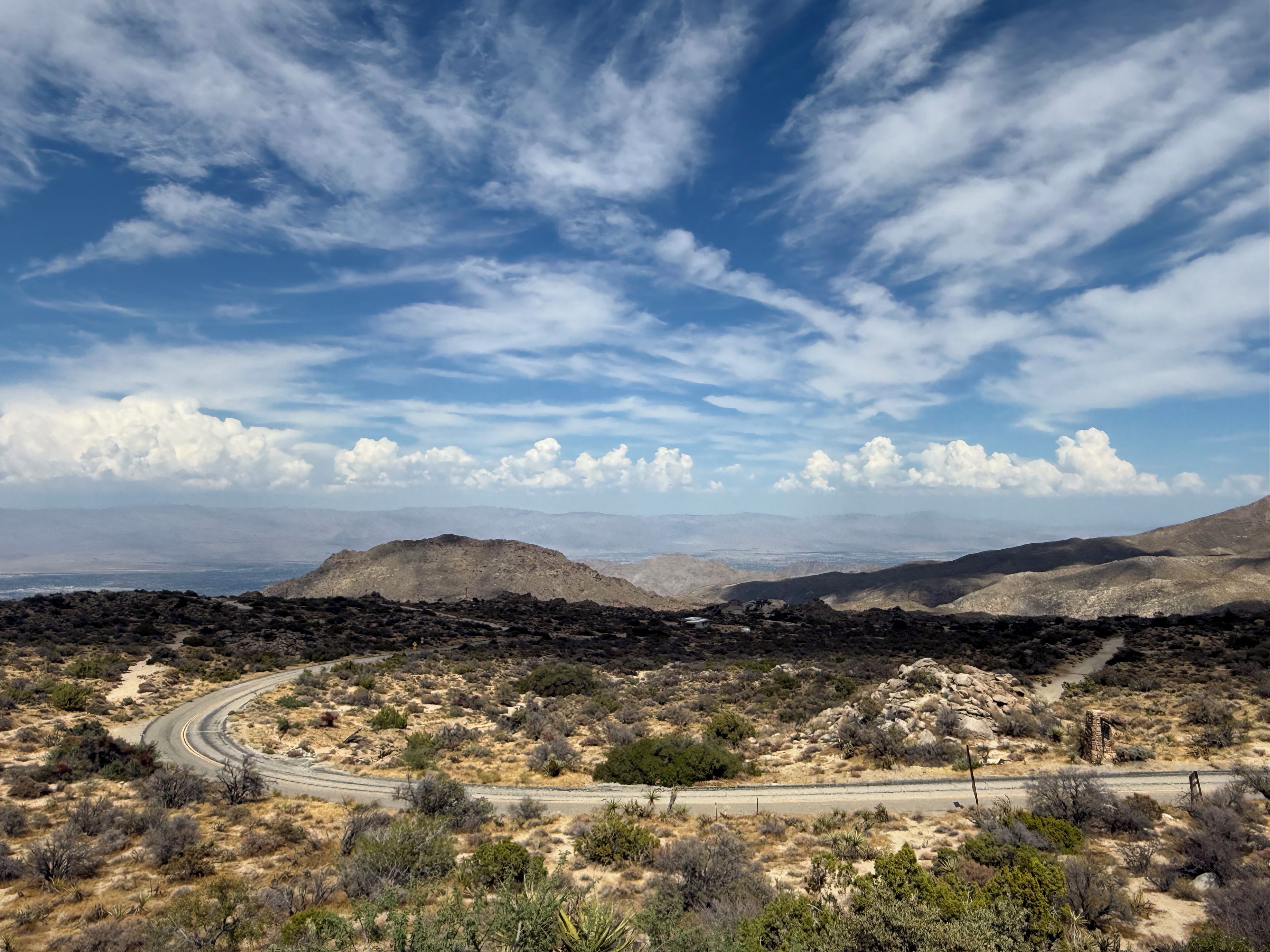 A desert landscape with a winding road, sparse vegetation, and distant mountains under a partly cloudy sky.