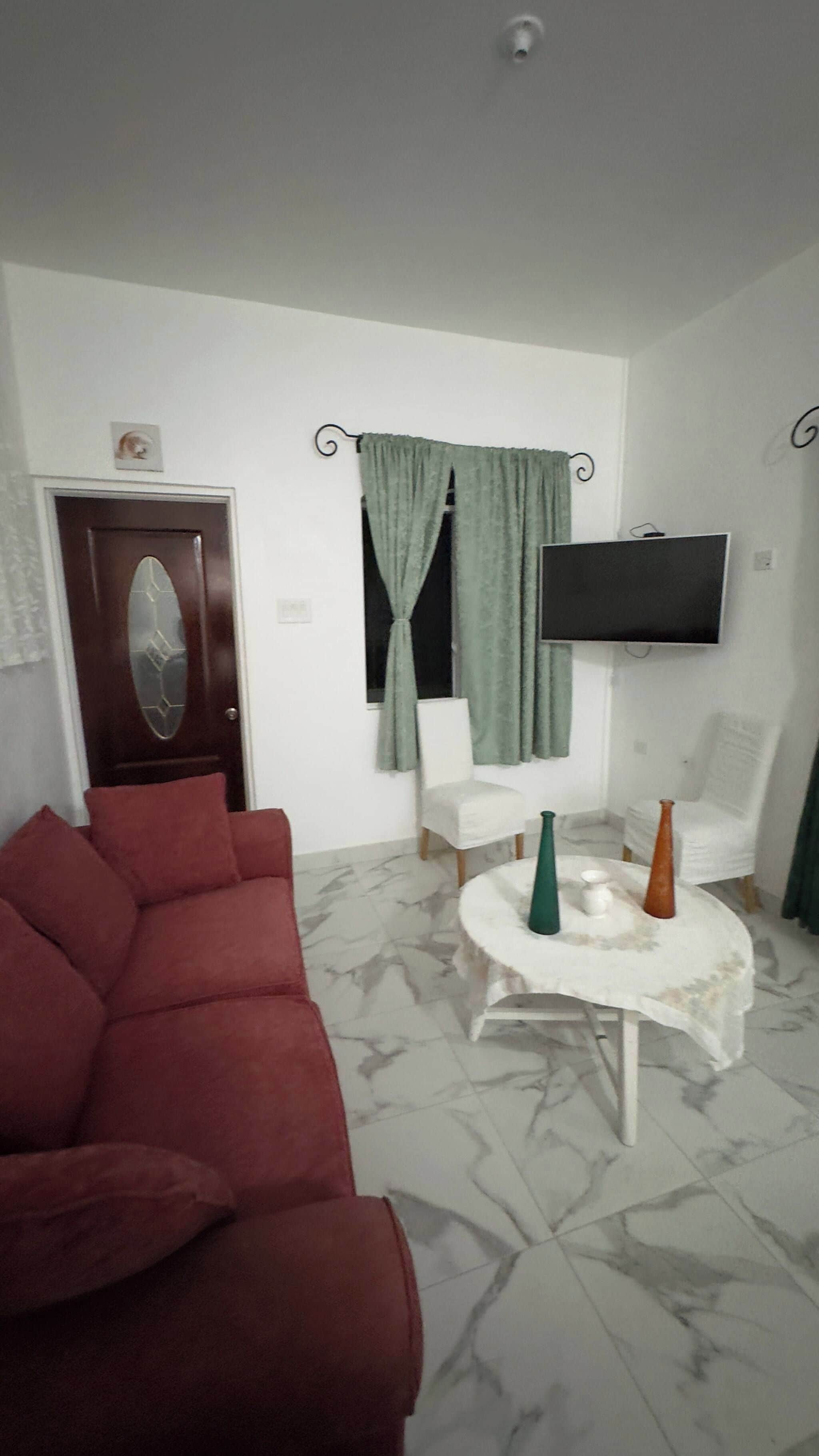 A living room with a red sofa, a white armchair, a small round table with decorative vases, a flat-screen TV mounted on the wall, and a door with a geometric glass panel.