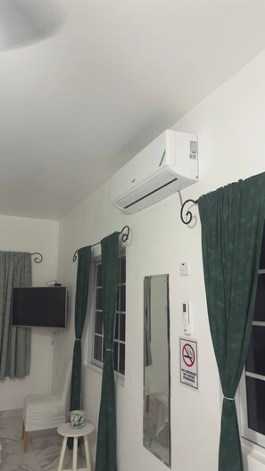 A room with a mounted air conditioner, a television on the wall, a small table with a cup, and a no-smoking sign. - Studio Flat Finished (Tollgate Clarendon Park)