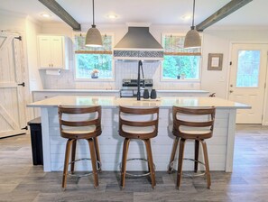 A kitchen with a large island, wooden bar stools, pendant lights, and a view of the outdoors. - Cozy Carolina Farmhouse • Near Crowders Mtn & Belmont (Gastonia)