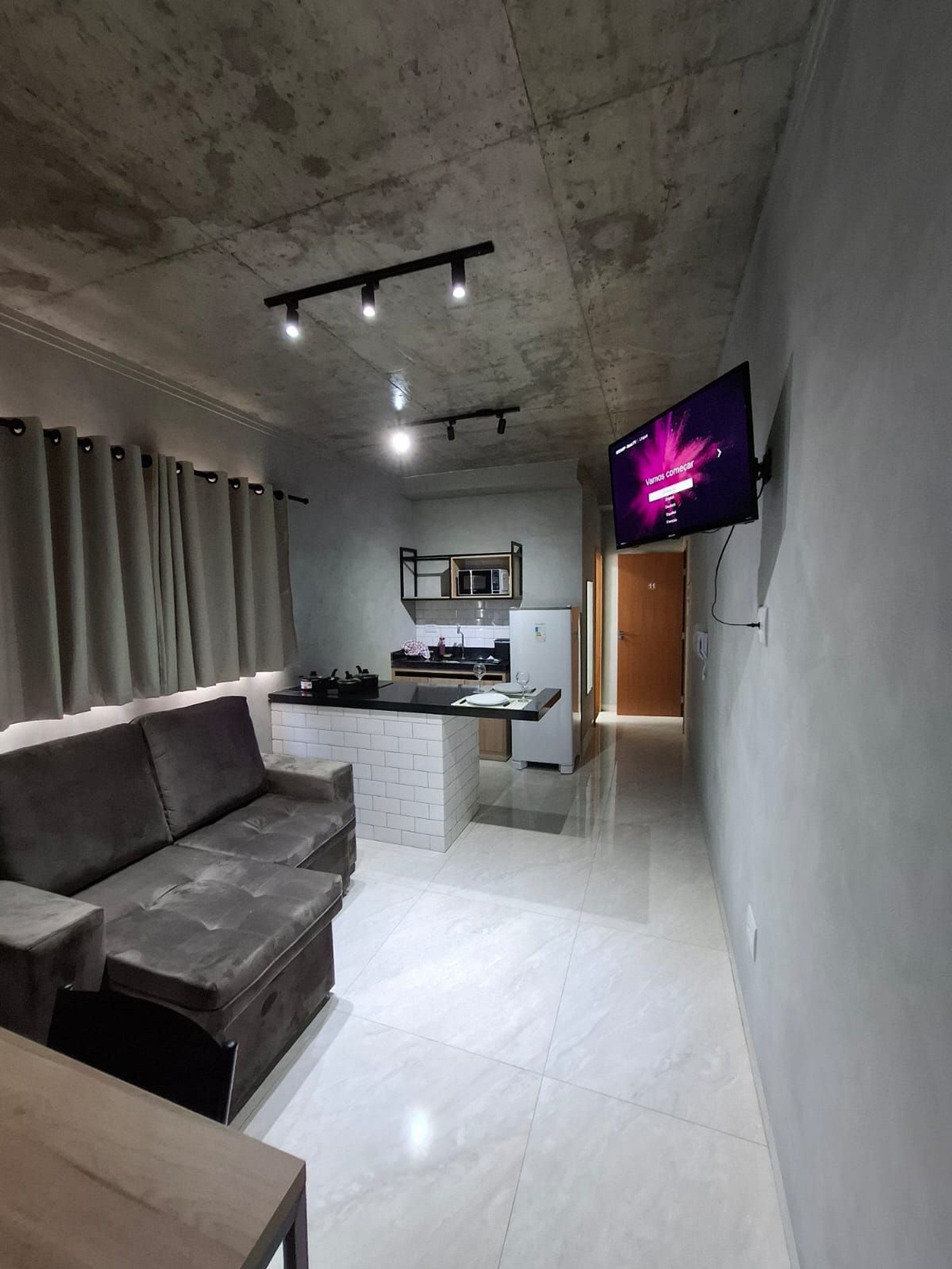 A modern living room with a sofa, a flat-screen TV mounted on the wall, and a kitchen area with a microwave and refrigerator.
