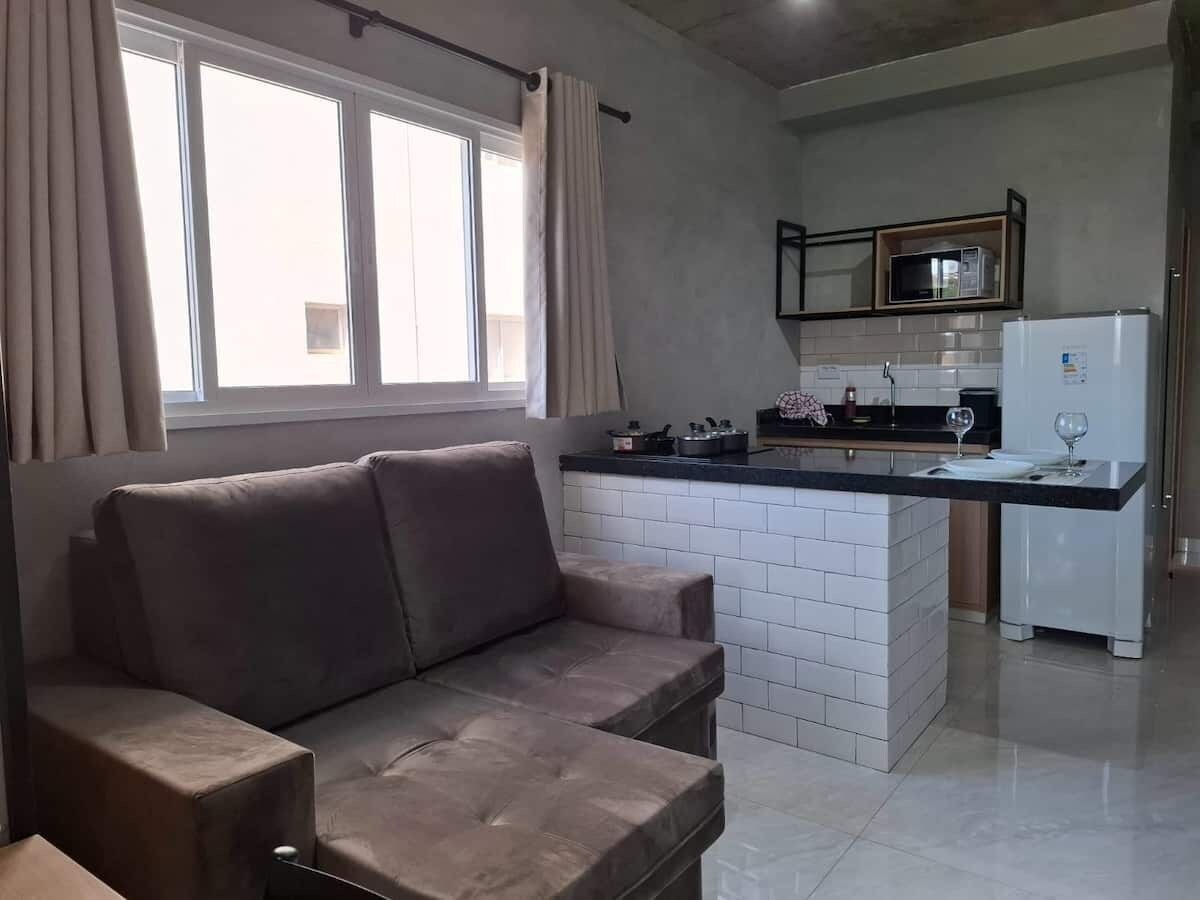A modern living room with a sofa, a kitchen area with a microwave and refrigerator, and a window with curtains.