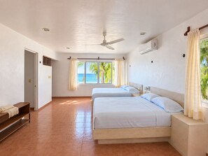 Habitación de hotel con dos camas, ventilador de techo, aire acondicionado, ventana con cortinas y vista al océano.