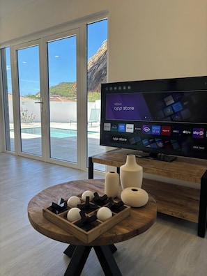 Een moderne woonkamer met een grote flatscreen-tv waarop een app-store interface te zien is, een houten salontafel met decoratieve objecten en uitzicht op een zwembad door glazen deuren.