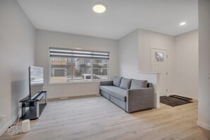 Ein modernes Wohnzimmer mit einem grauen Sofa, einem Fernseher auf einem Lowboard, einem großen Fenster mit Jalousien und einer Tür mit einem kleinen Bild.