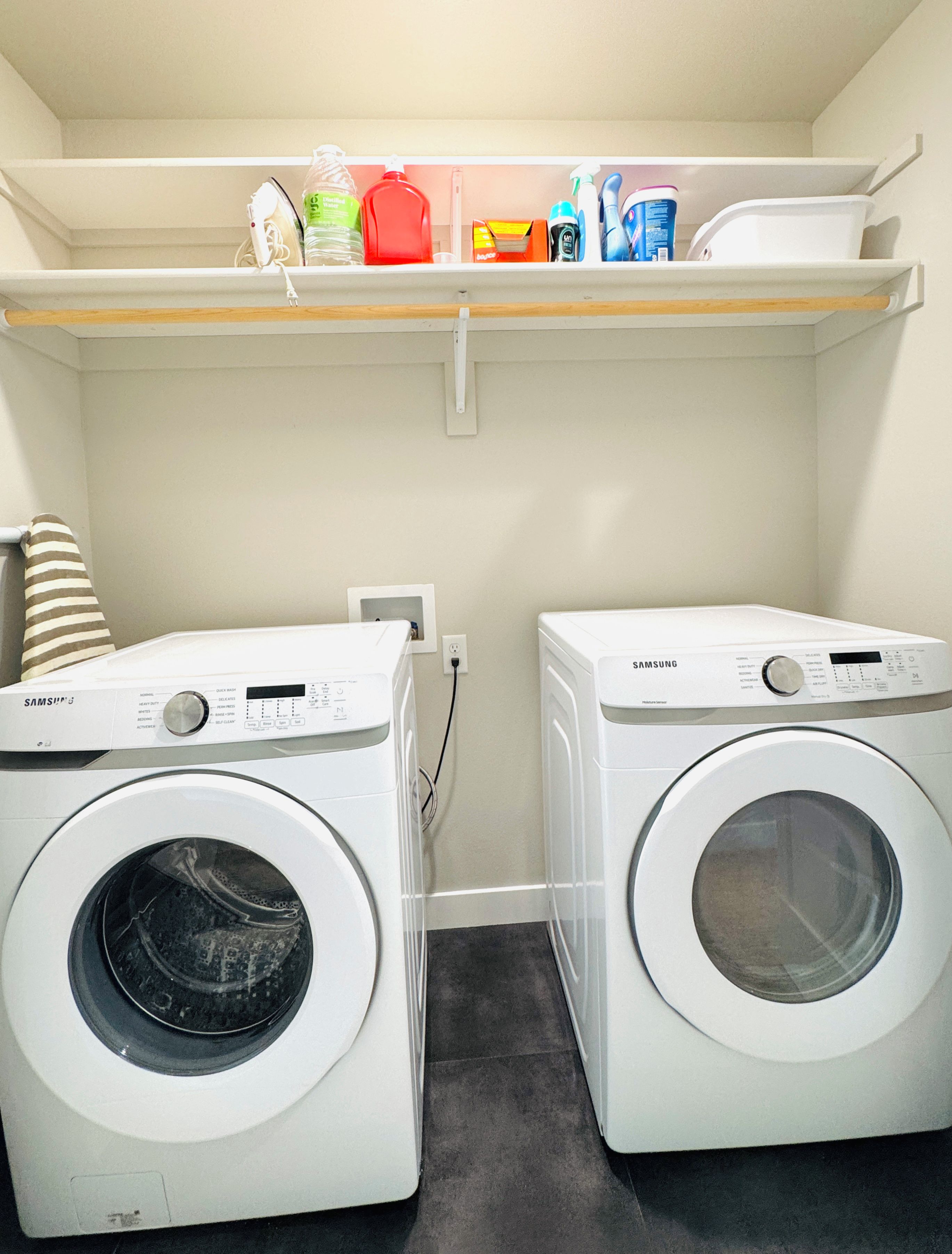 A laundry room with two Samsung washing machines, a shelf above with various cleaning supplies, and a striped towel on the left.