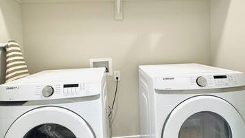 A laundry room with two Samsung washing machines, a shelf above with various cleaning supplies, and a striped towel on the left.